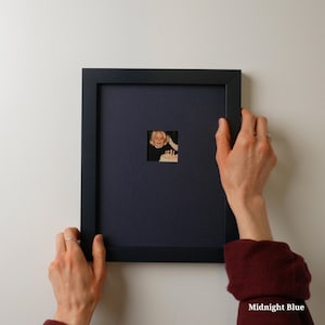 Op de afbeelding: Een zwarte fotolijst met een nachtblauwe passe-partout en een kleine foto. De lijst wordt door twee handen tegen een witte muur gehouden. De foto toont een lachende persoon die een taart vasthoudt.