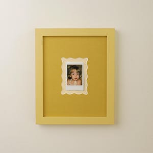 May include: A yellow framed picture with a small photo of a child. The frame is a solid yellow color, and the photo is centered within a scalloped-edge opening. The photo is a small Polaroid-style print.