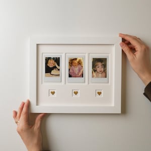 May include: A white rectangular picture frame featuring three vertical slots for square photos, each containing a different image of a child. Below each photo slot is a small square cutout containing a single gold-colored heart embellishment.