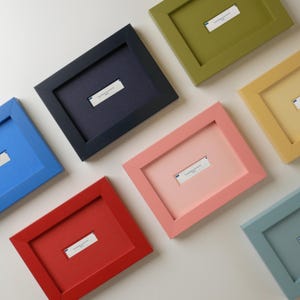 May include: A collection of rectangular picture frames in various solid colours including navy blue, olive green, bright red, coral pink, mustard yellow, sky blue, and muted teal. Each frame features a white rectangular label with small text.