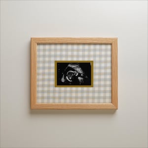 May include: A rectangular light wood frame containing a black and white ultrasound scan of a fetus. The scan is centered on a light gray and white gingham patterned background and is bordered by a thin, solid gold-colored rectangular mat.
