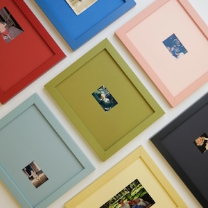 May include: A collection of seven rectangular picture frames in various solid colours including red, blue, olive green, light pink, light blue, yellow, and black. Each frame features a mount and contains a small photograph in the centre.