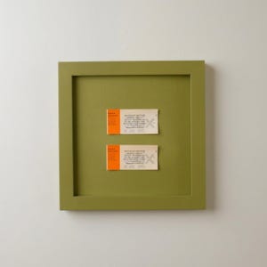 May include: A square olive green shadow box frame containing two rectangular tickets from the Dayton Art Institute. Each ticket features a bright orange vertical stripe on the left side, white text, and a faint gray X mark printed on the right side.