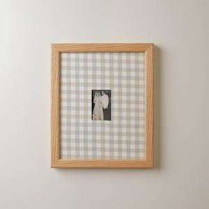 Pu&ograve; includere: Una fotografia incorniciata con un'immagine in bianco e nero di una coppia. La cornice &egrave; in legno chiaro e lo sfondo &egrave; un motivo a scacchi blu, grigio e bianco.