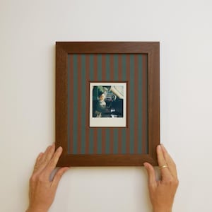 May include: A rectangular dark brown wooden picture frame featuring a dark green and brown vertical striped matting. The frame contains a small square photograph of a person sitting in the driver seat of a vehicle, captured in a bright outdoor setting.