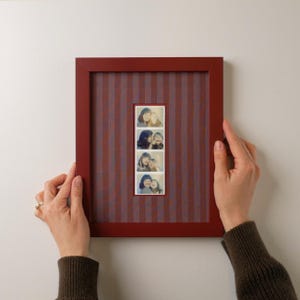 May include: A rectangular red picture frame with a vertical striped background and four small photos. The frame is being held by two hands, showcasing a simple yet elegant design for displaying cherished memories.