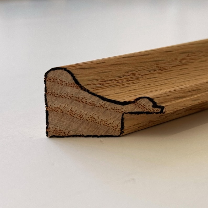 Puede incluir: Primer plano de una secci&oacute;n de moldura de madera sin terminar. La madera tiene una veta natural y es de color marr&oacute;n claro. El borde tiene una forma con un perfil curvo y un contorno negro, que revela la secci&oacute;n transversal de la madera.