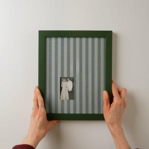 May include: A rectangular picture frame with a dark green border and a light gray and blue vertical striped matting. The center of the frame features a small, black and white photograph of two people standing together in formal attire.