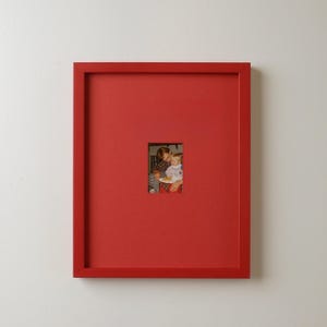 May include: A red picture frame with a small photograph in the center. The photograph shows a person holding a child. The frame is a solid, vibrant red color.