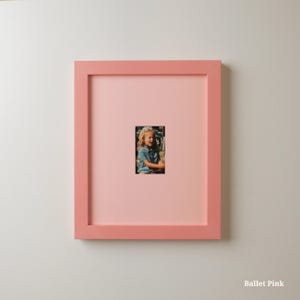 Op de afbeelding: Een vierkante, balletroze fotolijst met een kleine foto van een kind. De lijst is effen zachtroze van kleur en de foto is gecentreerd in een roze passe-partout. De tekst "Ballet Pink" staat rechtsonder.