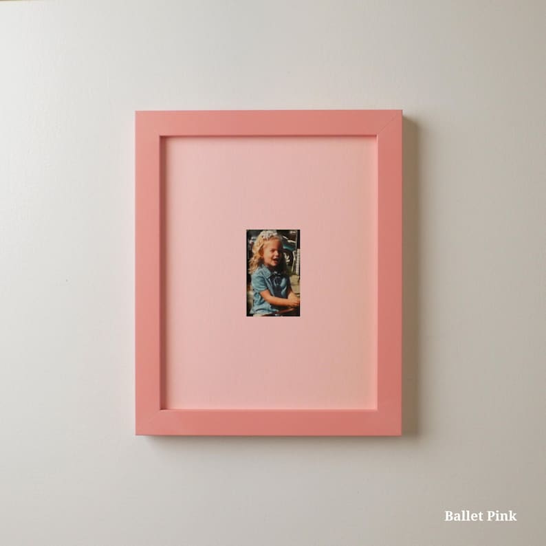 Op de afbeelding: Een vierkante, balletroze fotolijst met een kleine foto van een kind. De lijst is effen zachtroze van kleur en de foto is gecentreerd in een roze passe-partout. De tekst "Ballet Pink" staat rechtsonder.