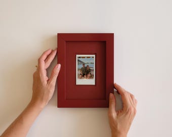 Könnte beinhalten: Ein tiefroter, rechteckiger Bilderrahmen mit einem mittig platzierten, kleineren Einsatzfenster, das ein quadratisches Foto von zwei Personen im Freien enthält. Der Rahmen hat eine glatte, matte Oberfläche und einen breiten Rand.