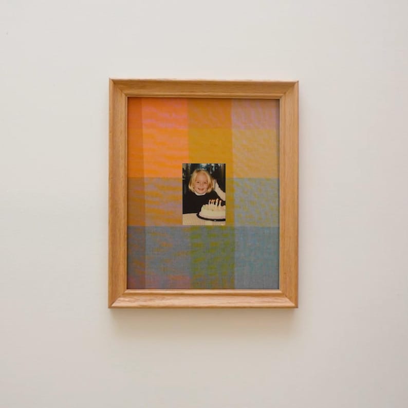 Puede incluir: Una obra de arte enmarcada con una peque&ntilde;a fotograf&iacute;a de una persona con una tarta de cumplea&ntilde;os, centrada en un colorido patr&oacute;n de cuadros. El marco es de madera clara, y el fondo de cuadros tiene cuadrados naranjas, amarillos, azules y verdes.