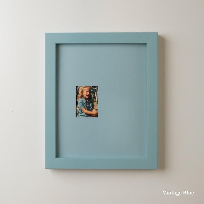 Op de afbeelding: Een vierkante, vintage blauwe fotolijst met een kleine foto van een kind. De lijst heeft een effen, lichtblauwe kleur en de foto is gecentreerd in de lijst. De tekst "Vintage Blue" staat rechtsonder.