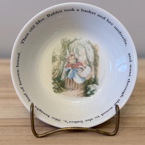 May include: White ceramic bowl with a colorful illustration of a rabbit in a forest scene. The bowl has text around the rim that reads, "Then old Mrs. Rabbit took a basket and her umbrella, and went through the wood to the baker's. She bought a loaf of brown bread."