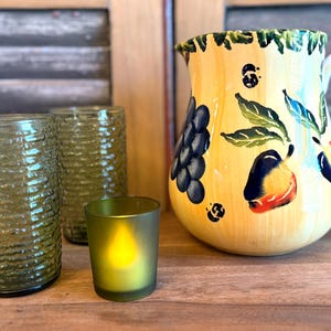 May include: A yellow ceramic pitcher with a white handle, decorated with painted fruit and leaves. Two textured green glasses and a small green candle holder are in front of the pitcher. The items are on a wooden surface.