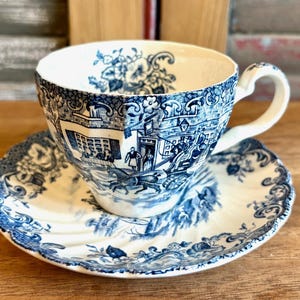 May include: A vintage teacup and saucer set. The white ceramic cup and saucer feature a blue transferware design depicting a rural scene with figures and a horse-drawn carriage. The cup has a curved handle and the saucer has a scalloped edge.