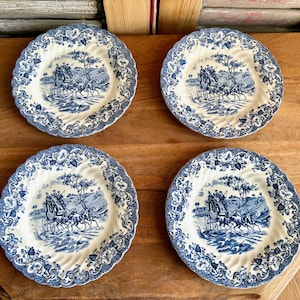 May include: Four blue and white decorative plates with a scalloped edge. Each plate features a detailed landscape scene with a horse-drawn carriage. The plates are arranged on a wooden surface.
