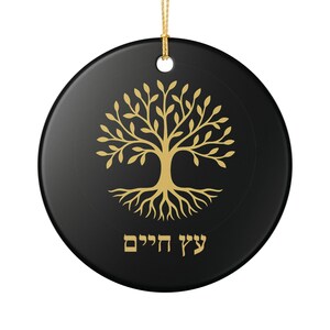 May include: A round, black ornament with a gold-colored Tree of Life design. The tree has a detailed trunk, branches, and roots. The text "עץ חיים" is written in gold at the bottom. A gold string is attached for hanging.