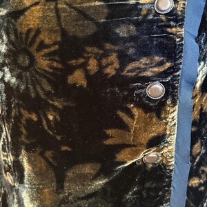 May include: Close-up of a dark velvet fabric with a floral pattern in shades of brown and gold. The fabric is detailed with antique-style buttons and a blue satin trim, suggesting a vintage garment.