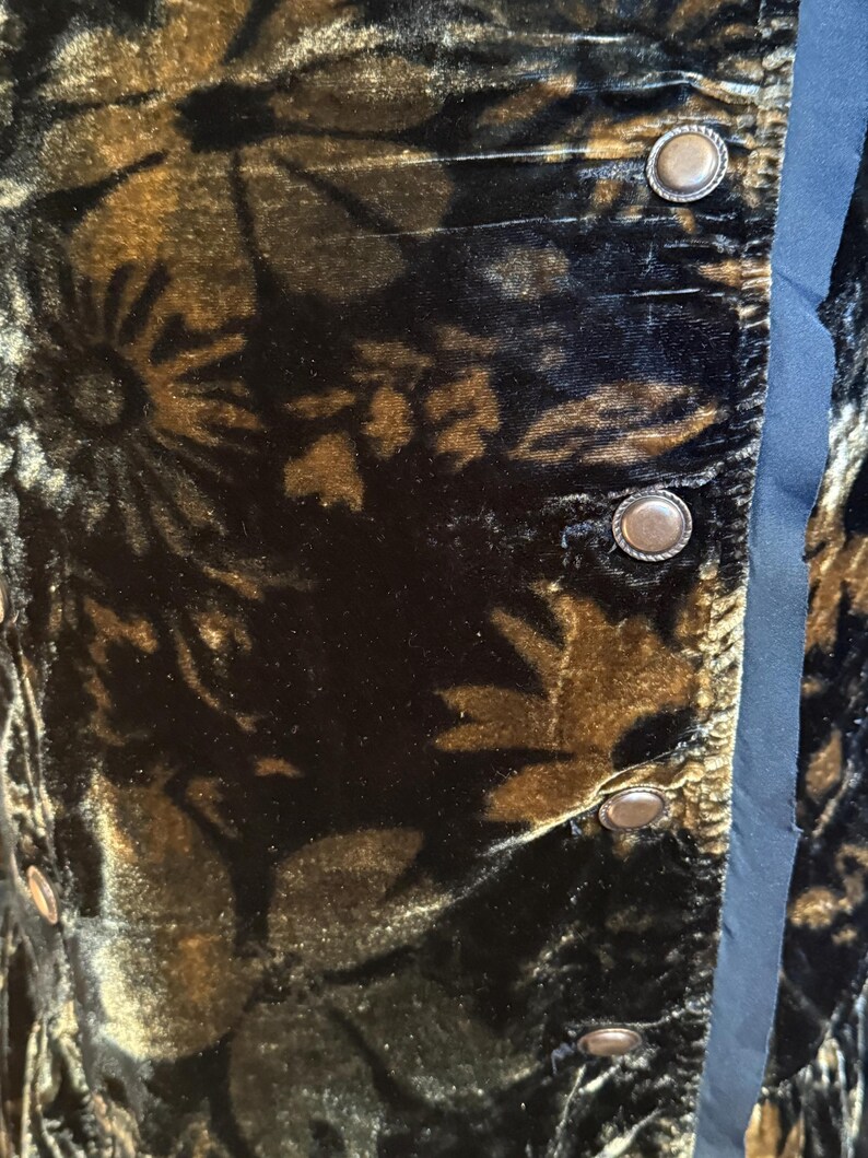 May include: Close-up of a dark velvet fabric with a floral pattern in shades of brown and gold. The fabric is detailed with antique-style buttons and a blue satin trim, suggesting a vintage garment.