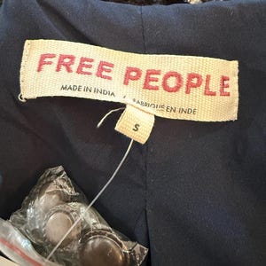 May include: Close-up of a navy blue garment with a tag that reads "FREE PEOPLE" in red letters. The tag also says "MADE IN INDIA" and "FABRIQUE EN INDE". A size "S" tag is attached. Several bottle caps are visible in a plastic bag.