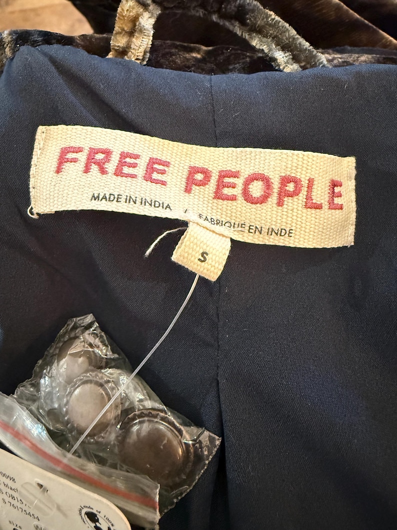 May include: Close-up of a navy blue garment with a tag that reads "FREE PEOPLE" in red letters. The tag also says "MADE IN INDIA" and "FABRIQUE EN INDE". A size "S" tag is attached. Several bottle caps are visible in a plastic bag.