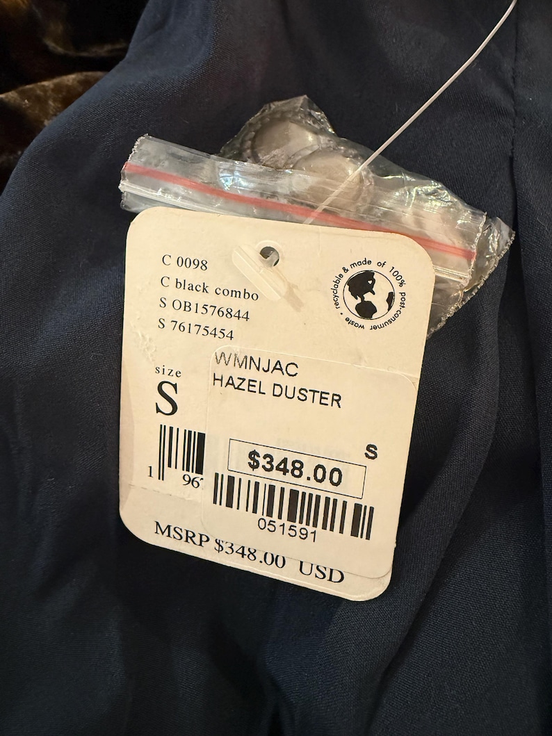 May include: A tag for a Hazel Duster in black combo, size small. The tag includes the product code, size, and a price of $348.00. The tag is attached to a dark blue garment.