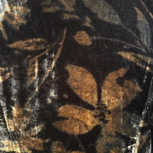 May include: Close-up of a black velvet fabric with a floral pattern in shades of brown and gold. The design features large, stylized flowers and leaves, creating a rich, textured appearance.
