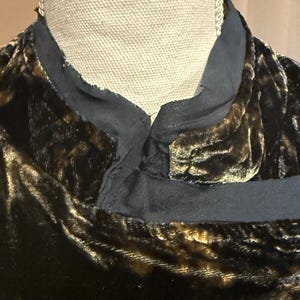 May include: Close-up of a dark brown velvet garment with a black collar. The velvet has a subtle sheen and a textured appearance. The collar is a contrasting black fabric. The garment is displayed on a mannequin.