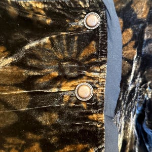 May include: Close-up of a section of a garment with a dark, patterned velvet fabric in shades of brown and black. The garment features three decorative, round buttons with a metallic finish and a black trim.
