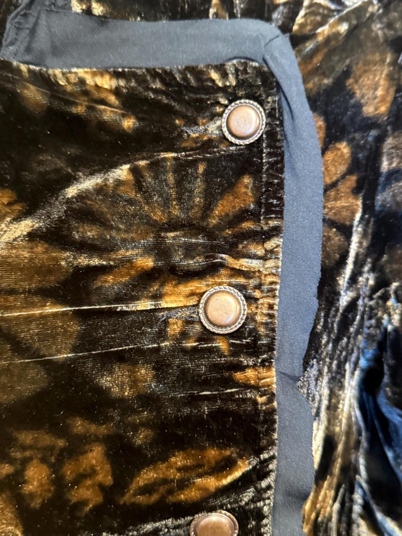 May include: Close-up of a section of a garment with a dark, patterned velvet fabric in shades of brown and black. The garment features three decorative, round buttons with a metallic finish and a black trim.