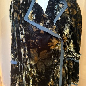 May include: A long, black velvet coat with a floral pattern in shades of gold and brown. The coat features a double-breasted design with silver buttons and a contrasting blue trim along the collar, lapels, and cuffs. The coat is displayed on a mannequin.