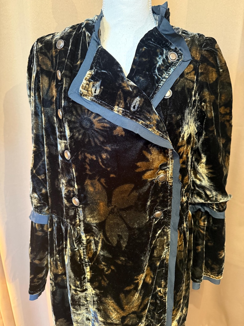 May include: A long, black velvet coat with a floral pattern in shades of gold and brown. The coat features a double-breasted design with silver buttons and a contrasting blue trim along the collar, lapels, and cuffs. The coat is displayed on a mannequin.