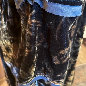 May include: Close-up of a dark, textured fabric with a velvet-like appearance. The fabric has a mix of black and brown tones, with a blue trim visible along the edge. The material appears soft and flowing.