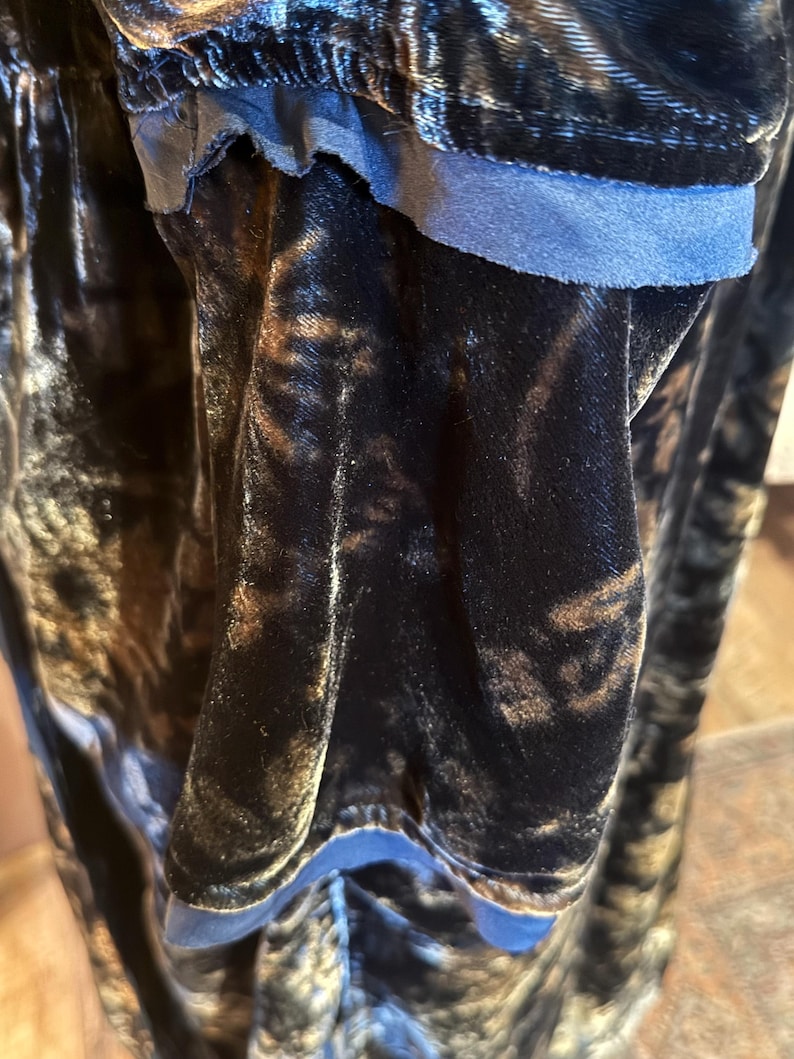 May include: Close-up of a dark, textured fabric with a velvet-like appearance. The fabric has a mix of black and brown tones, with a blue trim visible along the edge. The material appears soft and flowing.