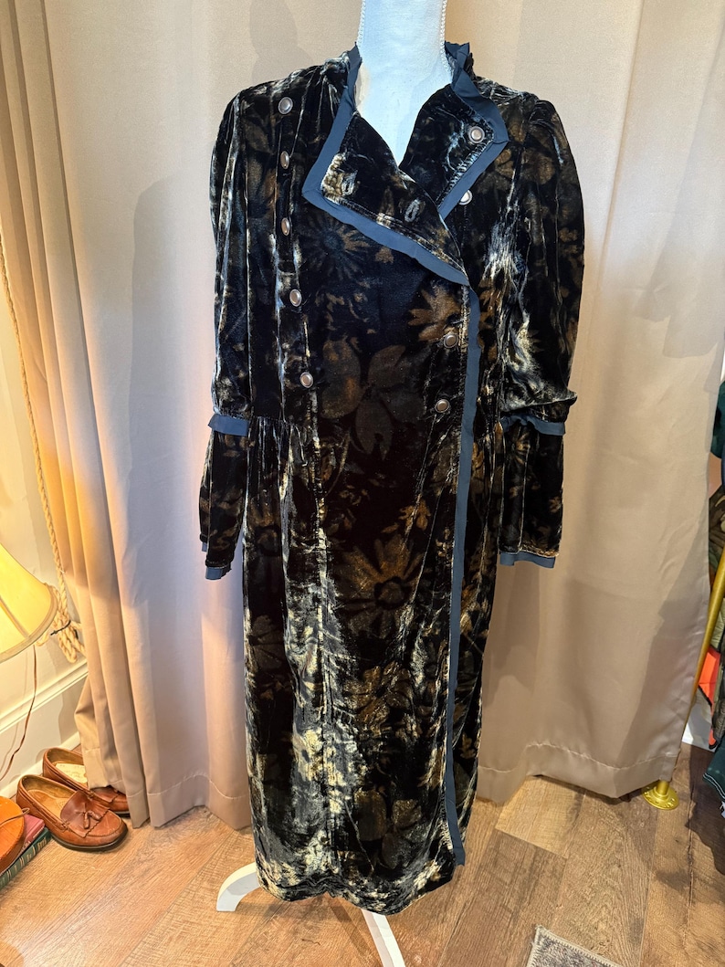 May include: A long, double-breasted coat in a dark floral pattern. The coat features a velvet texture, silver buttons, and blue trim along the collar, cuffs, and edges. The coat is displayed on a white mannequin.