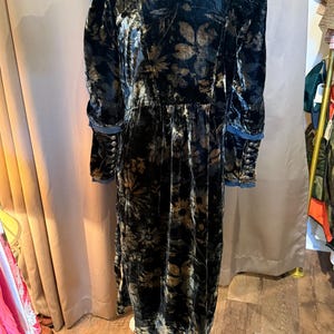 May include: A long, dark velvet dress with a floral pattern in shades of gold and brown. The dress features long sleeves with buttoned cuffs and a high collar. The dress is displayed on a white dress form.
