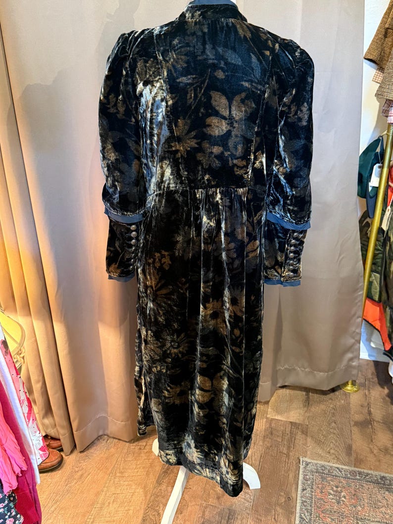 May include: A long, dark velvet dress with a floral pattern in shades of gold and brown. The dress features long sleeves with buttoned cuffs and a high collar. The dress is displayed on a white dress form.