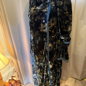 May include: A long, black velvet coat with a floral pattern in gold and olive green. The coat features a high collar, button closure, and a contrasting blue trim. The sleeves are long and flared.