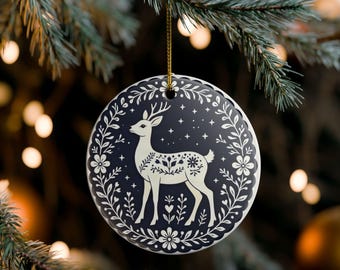May include: A round ceramic ornament featuring a dark navy blue background with a white illustration of a standing deer. The deer is surrounded by a circular border of white floral vines, leaves, and small stars. A gold string is attached to the top.