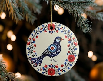 May include: A round, cream-colored ceramic ornament featuring a dark blue bird illustration at the center. The bird is surrounded by a circular wreath of red and blue floral patterns. A gold string is attached to the top for hanging purposes.