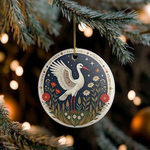 Può includere: Una decorazione rotonda in ceramica bianca con l'illustrazione di una cicogna bianca dalle ali spiegate, circondata da fiori rossi e bianchi su uno sfondo blu scuro stellato, dotata di un cordino dorato per appenderla.