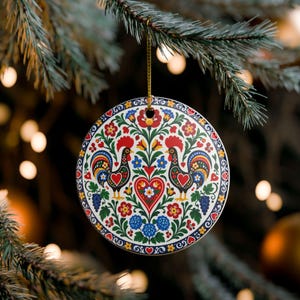 May include: A round ceramic ornament featuring a vibrant folk art design with two roosters facing a central heart, surrounded by colourful floral patterns in red, blue, yellow, and green, suspended by a thin gold cord from a pine tree branch.