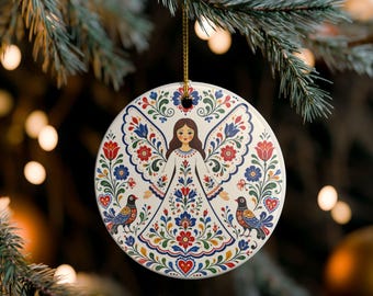 May include: A round white ceramic ornament featuring a central illustration of an angel with brown hair, surrounded by symmetrical floral patterns in red, blue, and green, with two birds at the bottom and a gold hanging string at the top.