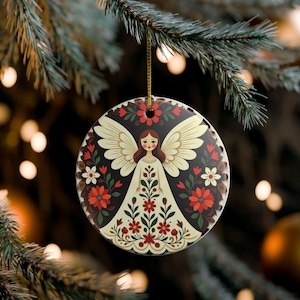 May include: A round ceramic ornament featuring a central illustration of an angel with cream-colored wings and a dress adorned with red and white floral patterns. The background is dark, accented by a symmetrical arrangement of red and white flowers.