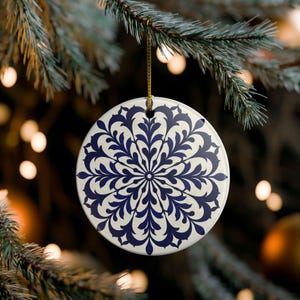 May include: A round, white ceramic ornament featuring a dark blue, symmetrical, radial floral pattern. The ornament is suspended by a thin gold string, showcasing a detailed, intricate design that radiates from the center toward the edges.