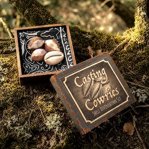 May include: A wooden box set for casting cowries, featuring a black and white bandana with four cowrie shells. The set includes a second box with the text "Casting Cowries Shell Divination Set" in gold lettering.