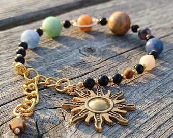 May include: A celestial-themed bracelet featuring a gold sun charm and a chain. The bracelet includes colorful beads representing planets, such as blue, green, orange, and brown, interspersed with black beads. The bracelet is set against a wooden surface.