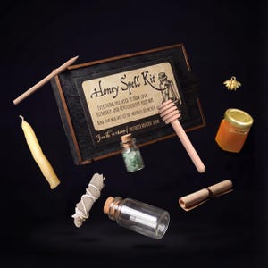 May include: A Honey Spell Kit with various components, including a wooden box, a candle, a honey dipper, a small jar of honey, a scroll, and a small bottle with green stones. The kit includes the text "Honey Spell Kit". A gold bee charm is also present.